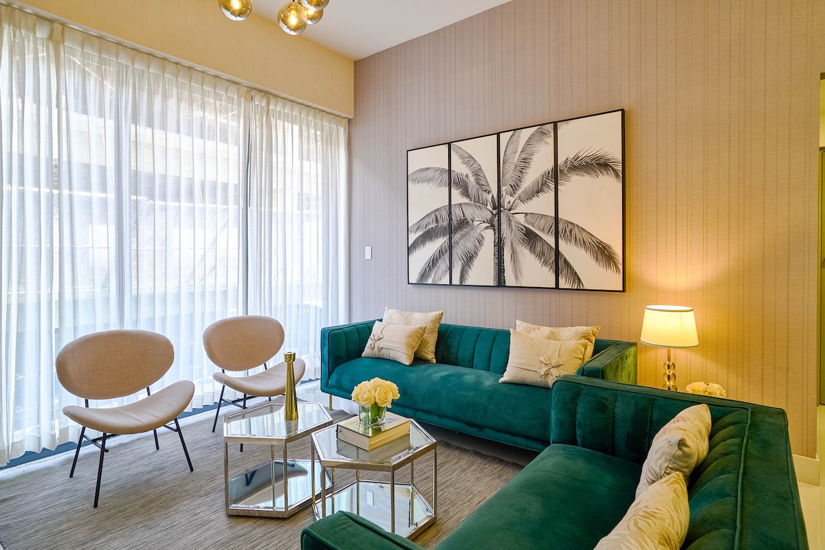 A modern living area features a green sectional sofa and two beige accent chairs. A glass coffee table sits at the center, adorned with flowers. Natural light filters through sheer curtains, highlighting the palm-themed wall art above the sofa.