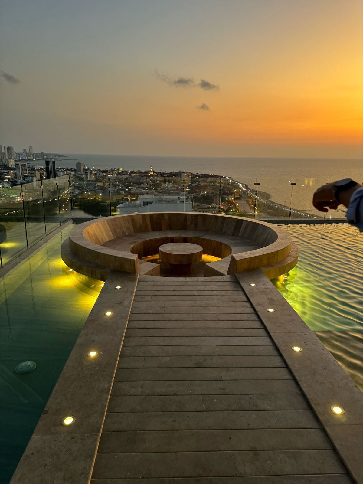 A circular hot tub is situated at the edge of a pool, offering sweeping views of the ocean and city skyline at sunset. Soft lighting illuminates the surrounding area, enhancing the tranquil setting as the sun sets over the horizon.