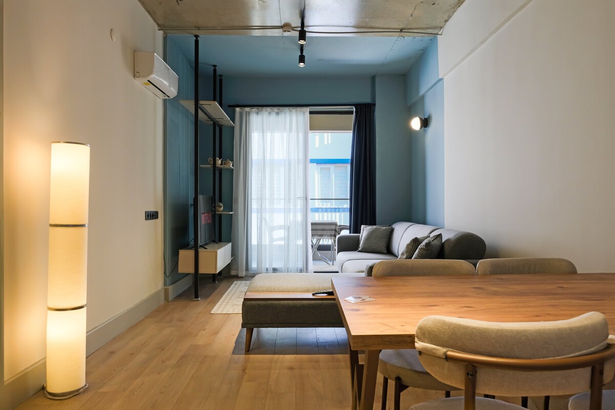 A modern living area features a light wooden dining table with several chairs and a comfortable gray sofa. A floor lamp stands beside the sofa, and large glass doors lead to a balcony, allowing natural light to fill the space.