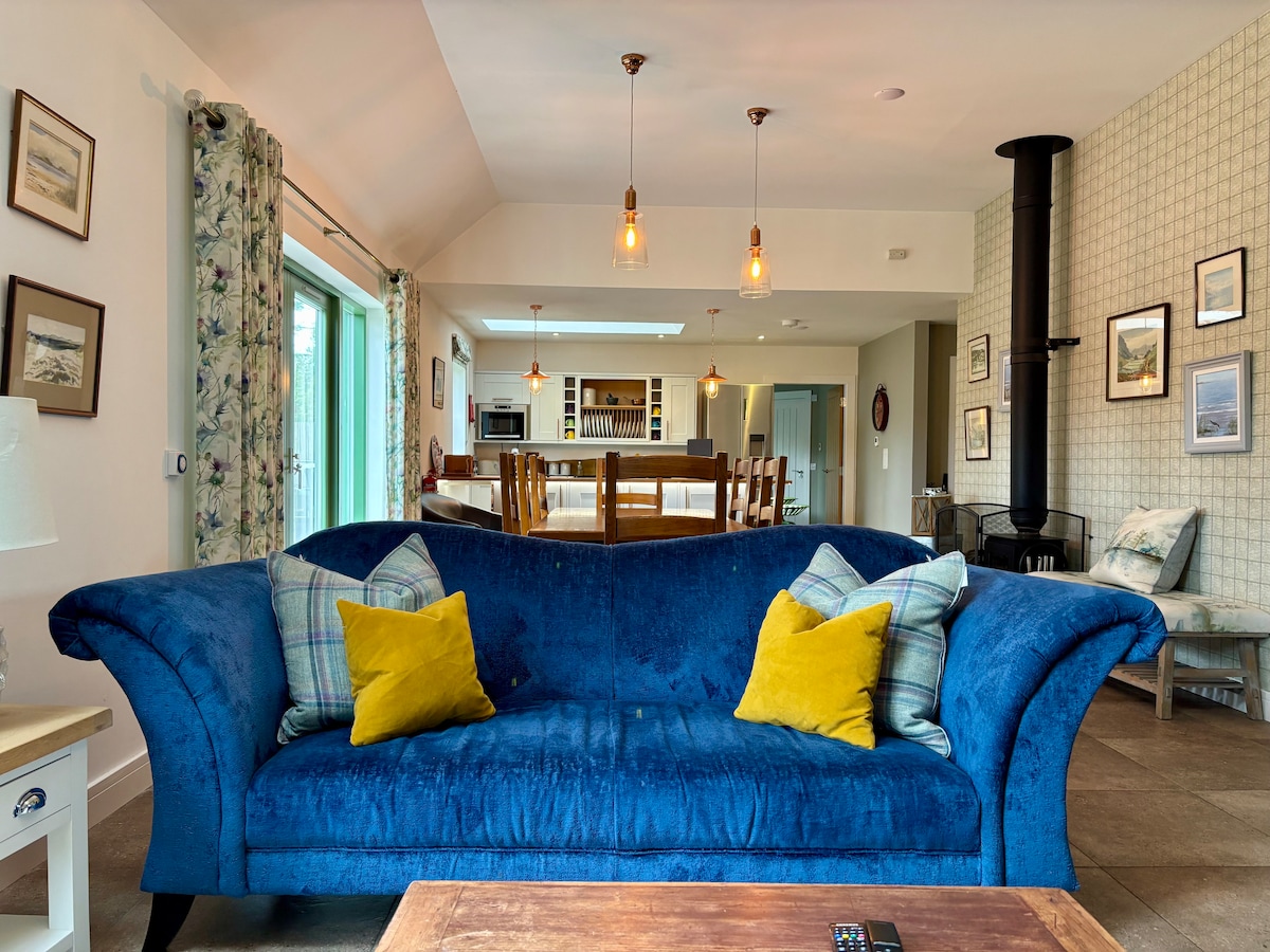 A spacious living area is furnished with a deep blue sofa adorned with yellow accent pillows. In the background, a well-lit kitchen area is visible, featuring a dining table and a modern fireplace. Natural light enters through large glass doors, enhancing the inviting space.
