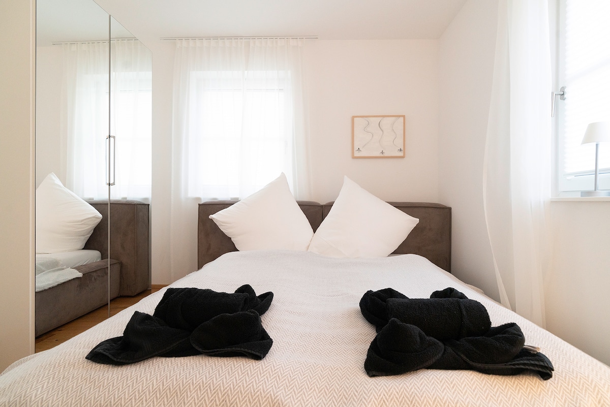 A spacious bed is centered in the room, adorned with two large pillows and neatly arranged dark towels. Natural light filters through sheer curtains, illuminating the neutral color scheme. A mirror doubling as a wardrobe stands against the wall, enhancing the airy feel of the space.