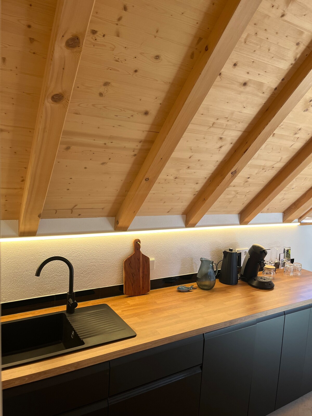 A modern kitchen area features a wooden countertop, enhanced by under-cabinet lighting. A sleek black sink and stylish faucet are positioned next to a wooden cutting board, with various kitchen appliances and glassware arranged thoughtfully on the counter.