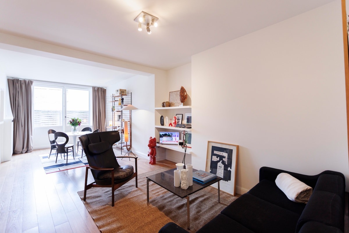 A spacious living area features a comfortable black sofa and a stylish coffee table. An accent chair is positioned nearby, with a well-lit floor lamp illuminating the space. Shelves display decor items, while large windows invite natural light and offer views of the outdoors.