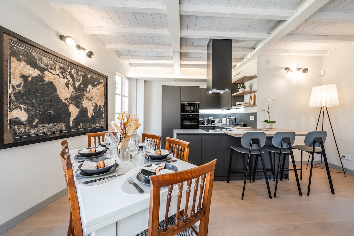 The dining area features a white table set with grey plates and glassware, surrounded by wooden chairs. A world map hangs on the wall, complementing the modern kitchen equipped with sleek black cabinetry and appliances. Natural light fills the space, enhancing its airy feel.