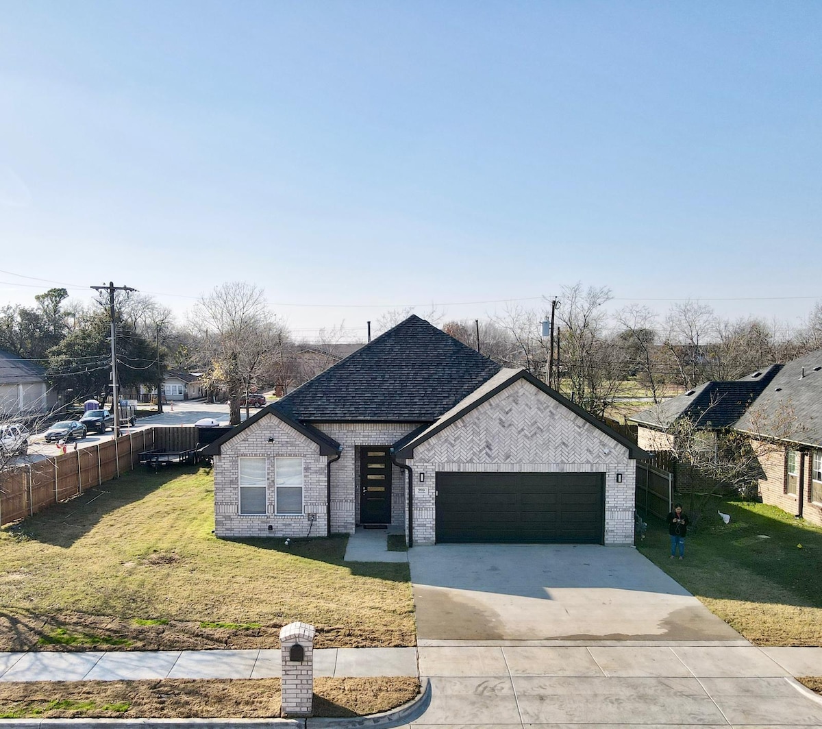 A modern single-story home with a brick exterior and a symmetrical design is displayed. A spacious driveway leads to a two-car garage. The yard is maintained with green grass, and surrounding trees can be seen in the background, signifying a suburban neighborhood.