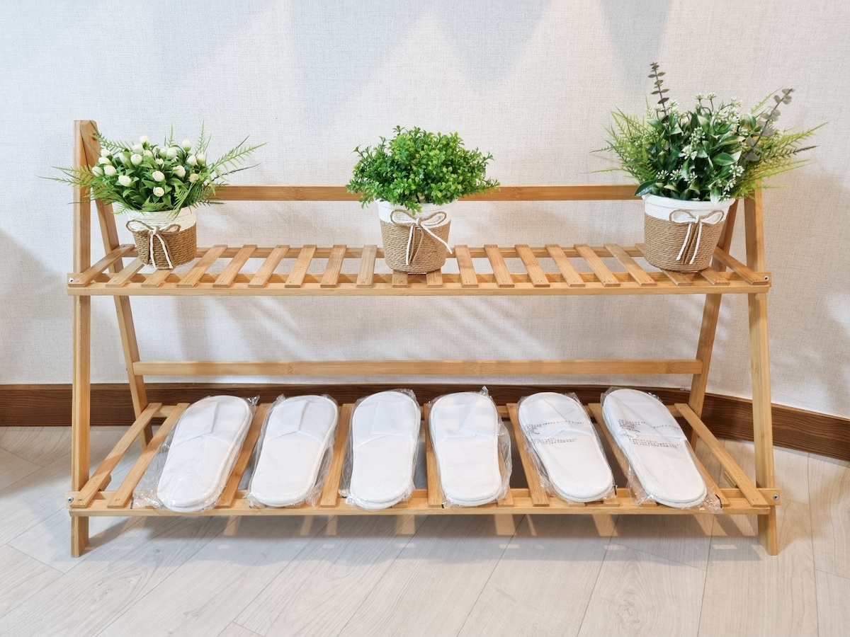 A wooden shoe rack is positioned against a wall, featuring two tiers. The top tier displays potted plants, while the bottom tier holds neatly arranged white slippers, each protected in plastic. The overall design conveys a welcoming touch to the accommodation.