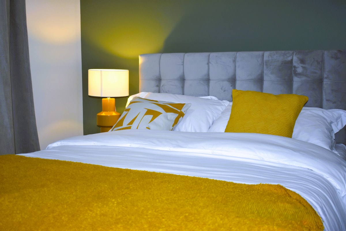 A cozy bedroom is highlighted by a plush bed with white linens and accent pillows. A vibrant yellow throw blanket drapes over the edge. Soft lighting from a bedside lamp creates a calming atmosphere against the dark green wall.
