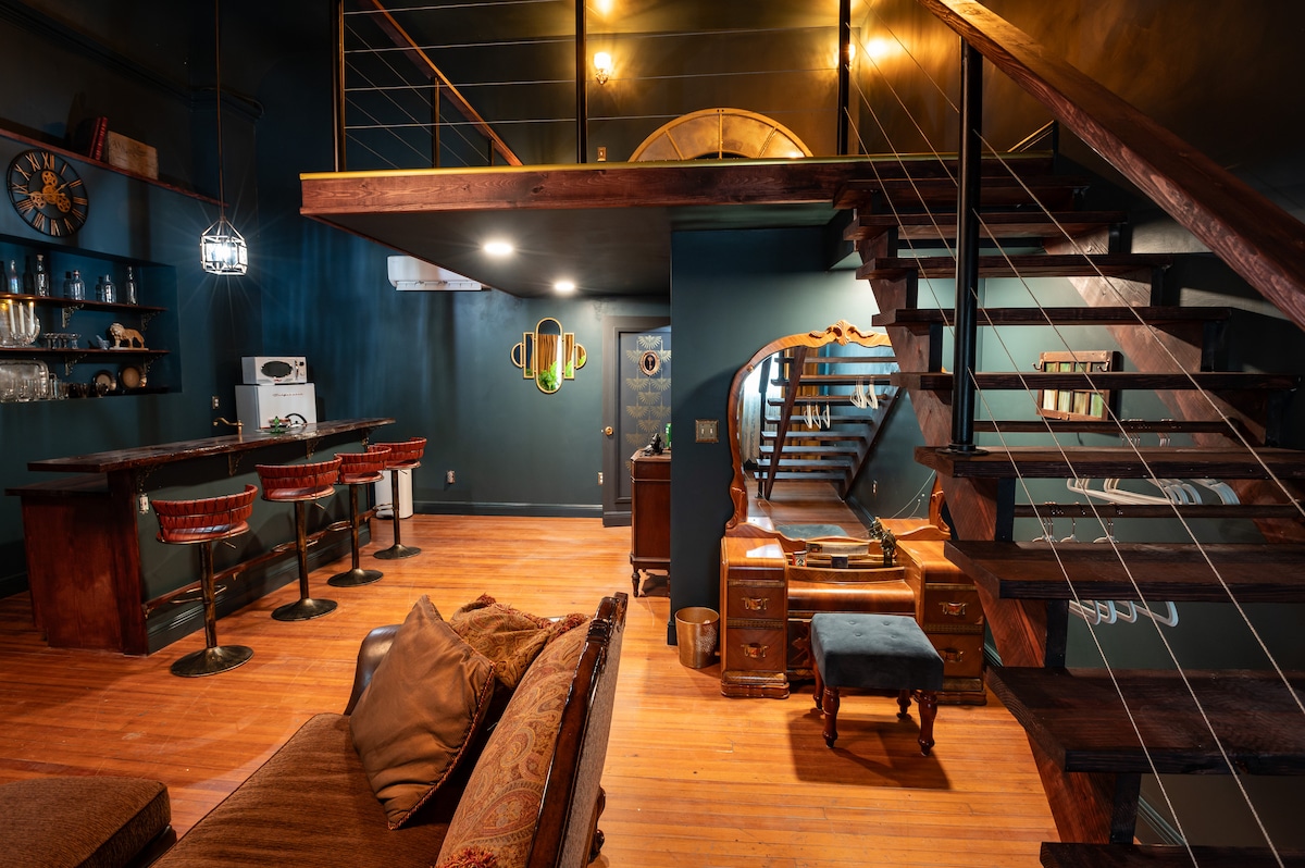 A cozy living area features rustic wood flooring and vintage furnishings, including a plush sofa with decorative pillows. A modern bar counter with tall chairs occupies one corner, while a mirrored vanity and staircase lead to the lofted bedroom above, creating a spacious and inviting atmosphere.