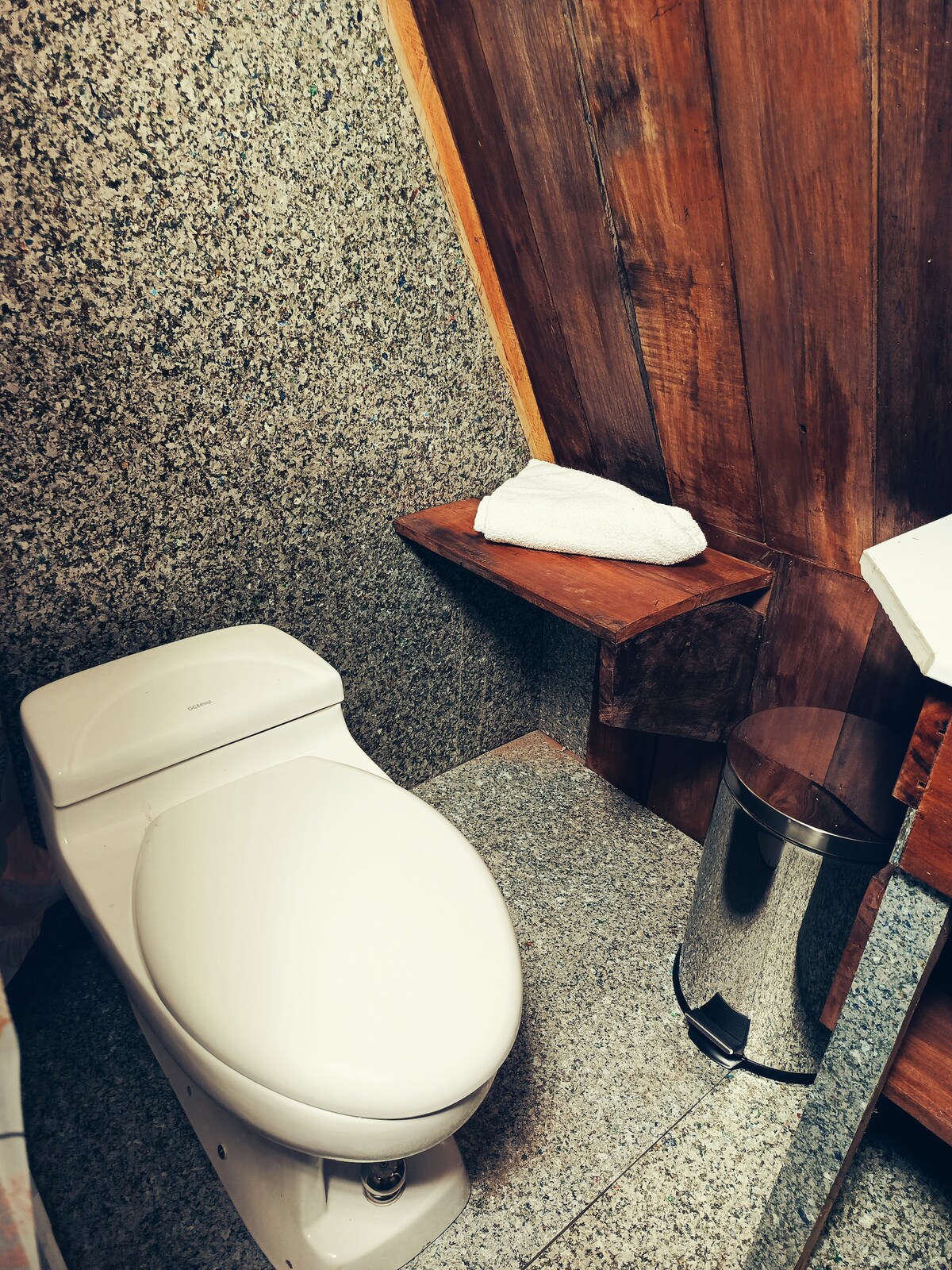 A clean and modern bathroom features a white toilet alongside a wooden shelf holding a rolled towel. A metallic trash can completes the space, which is accented by textured stone walls and a smooth countertop.
