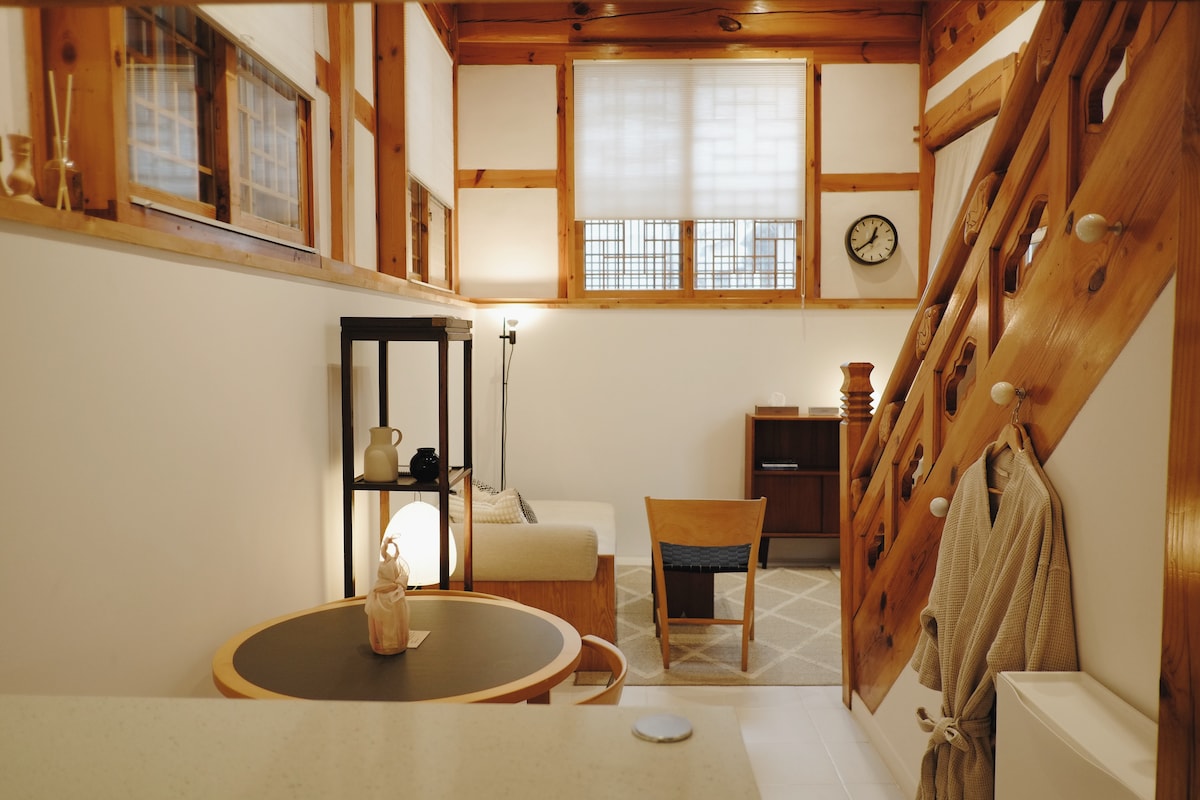 The interior showcases a blend of traditional and modern design elements. A circular table sits at the forefront, with a light fixture illuminating the space. A comfortable armchair and a small cabinet are arranged for relaxation. Natural wood beams and a wall clock add a rustic touch.