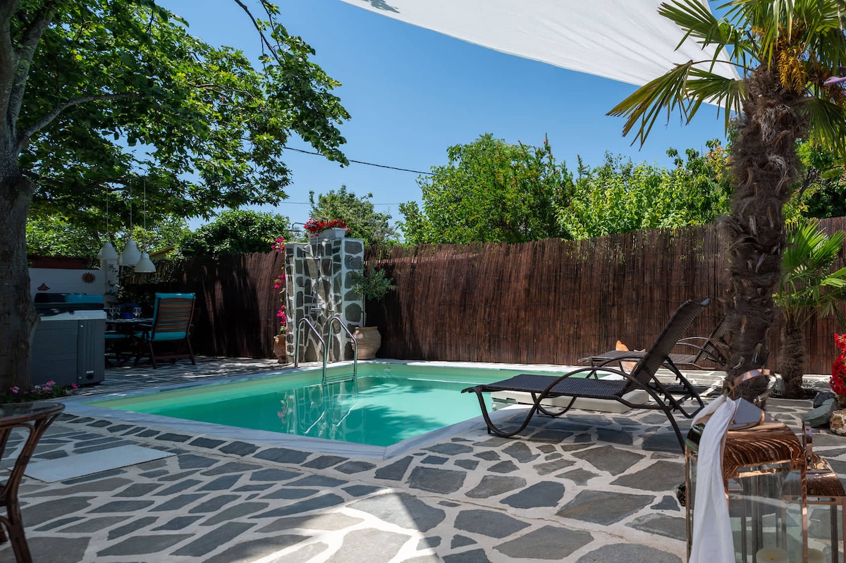 A serene outdoor area features a rectangular pool surrounded by natural stone flooring. Sun loungers are positioned nearby under a shaded area, with lush greenery providing privacy. Decorative plants and a wooden fence contribute to a peaceful atmosphere suitable for relaxation.