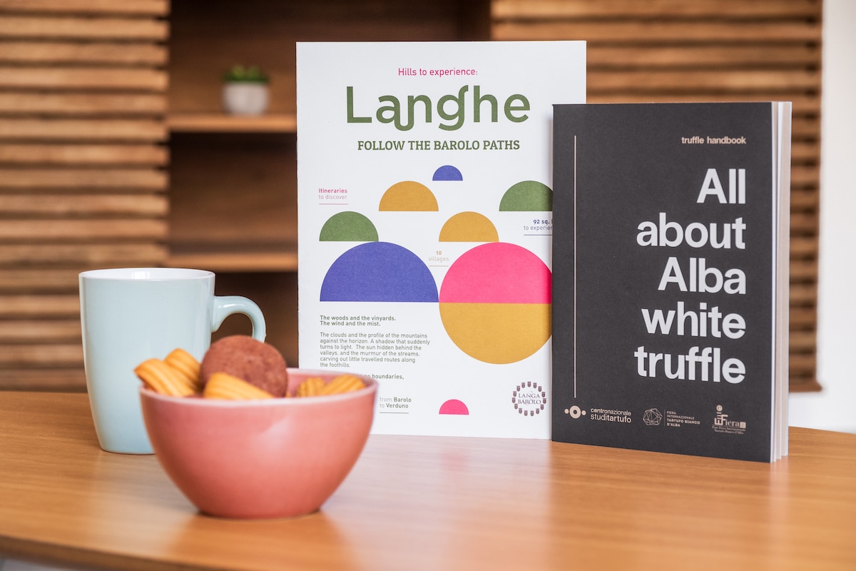 A pink bowl filled with cookies and pastries is positioned next to a light blue mug on a wooden tabletop. Informational booklets about Langhe and truffles are neatly placed upright, with vibrant colors and bold graphics, adding a welcoming touch to the cozy setting.