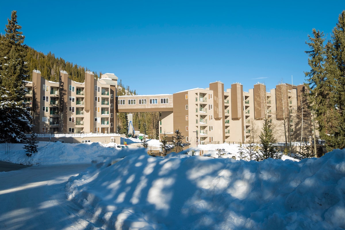The exterior of the condo complex is showcased against a clear blue sky, surrounded by snow-covered ground and evergreen trees. The building's modern architecture features various levels and large windows, offering a contemporary design that blends with the natural mountain environment.