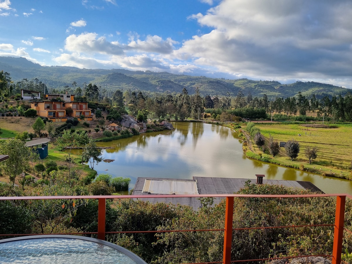 A serene view of a tranquil lake is framed by lush greenery and distant mountains. The landscape features scattered buildings along the shore and a bright sky with some clouds, enhancing the natural beauty of the surroundings.