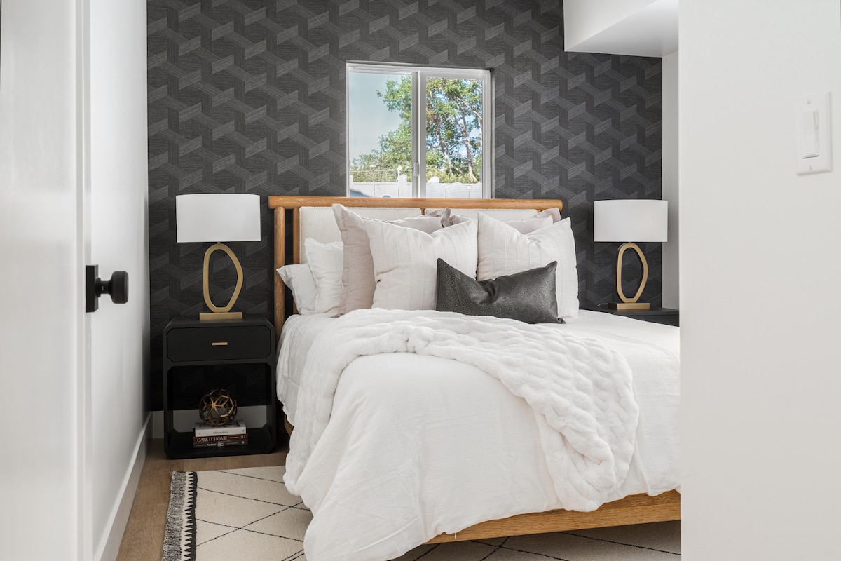 A cozy bedroom features a wooden bed with a layered white comforter and numerous pillows. Two bedside lamps sit on dark nightstands, providing soft lighting. A window allows natural light to enter, with a leafy view framed by modern geometric wallpaper.