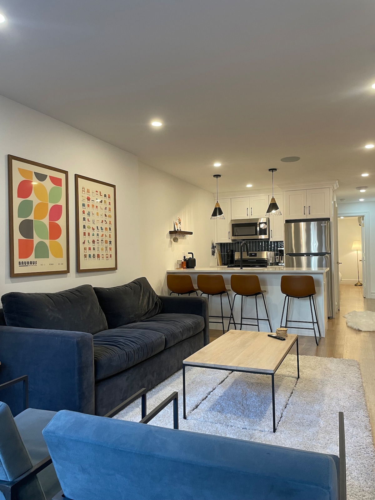 The living area features a comfortable dark sofa paired with two accent chairs, set against a wall adorned with framed colorful artwork. A light-colored coffee table sits atop a soft rug, while the open kitchen is equipped with modern appliances and bar seating for four.