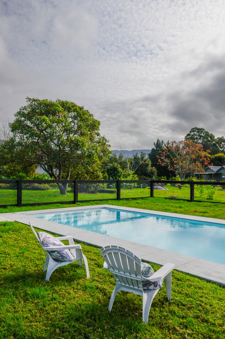 A swimming pool is framed by a well-maintained lawn and carefully placed white lounge chairs. The tranquil outdoor space is highlighted by surrounding greenery and trees, creating a serene environment. Soft clouds are visible in the sky, adding to the calm atmosphere.