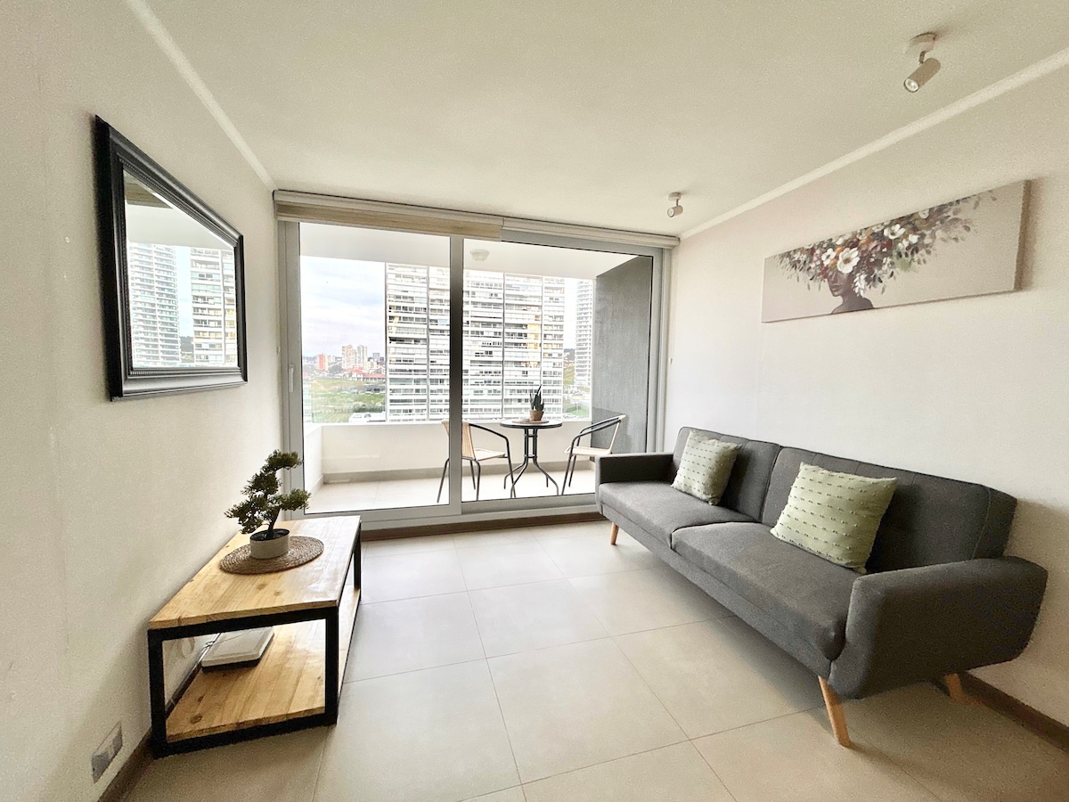 A modern living area features a sofa complemented by two decorative pillows. A wooden coffee table sits nearby, with a small indoor plant on top. Large glass doors provide a view of the cityscape and access to a balcony with seating for two.