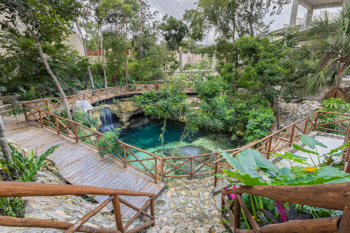 A serene cenote is surrounded by lush greenery and wooden bridges. The clear water reflects the surrounding flora and a gentle waterfall can be seen cascading into the pool, creating a peaceful natural setting.