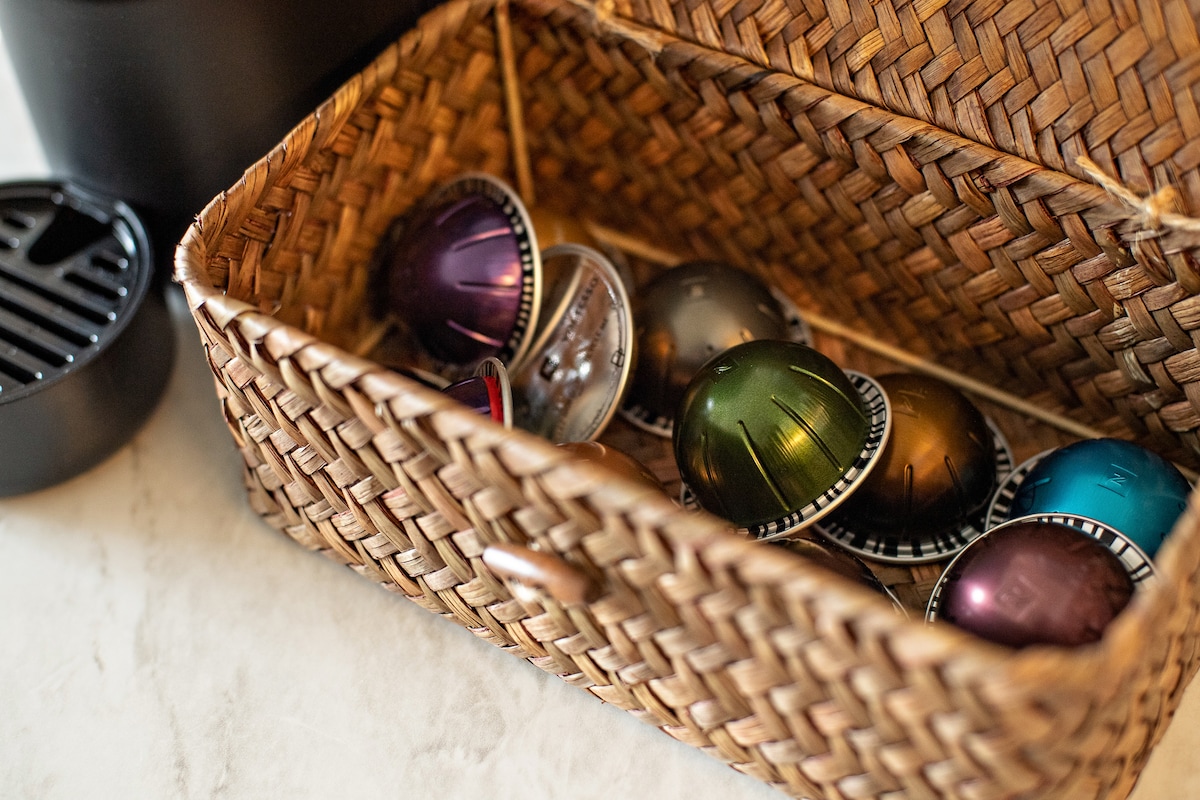 A woven basket is featured on a countertop, holding a variety of coffee capsules in multiple colors, including purple, green, and teal. A coffee machine stands nearby, enhancing the kitchen's inviting atmosphere.