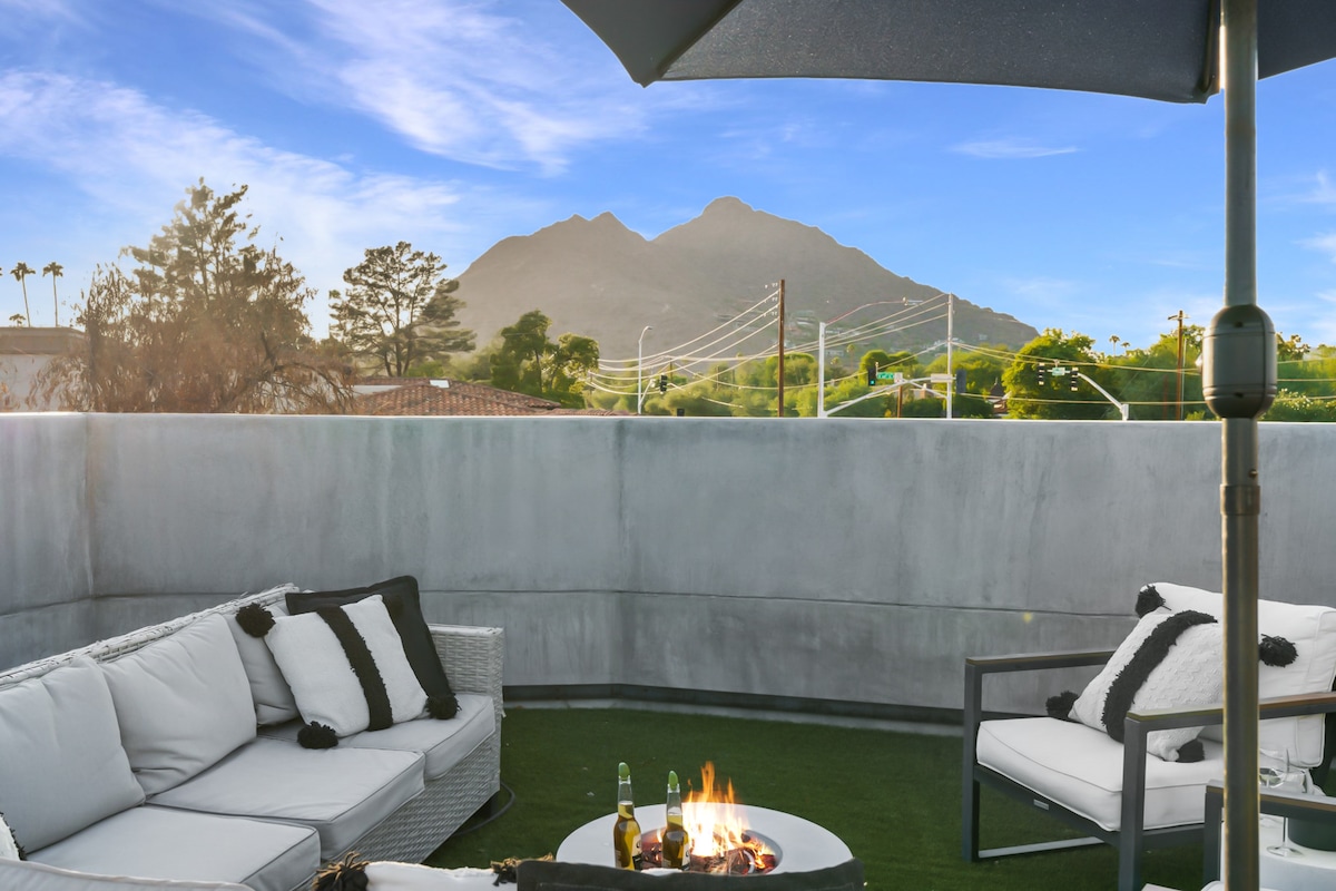 A rooftop lounge area is furnished with comfortable seating, featuring a table with a fire pit at its center. The backdrop includes a clear view of Camelback Mountain under a bright blue sky, surrounded by lush greenery.
