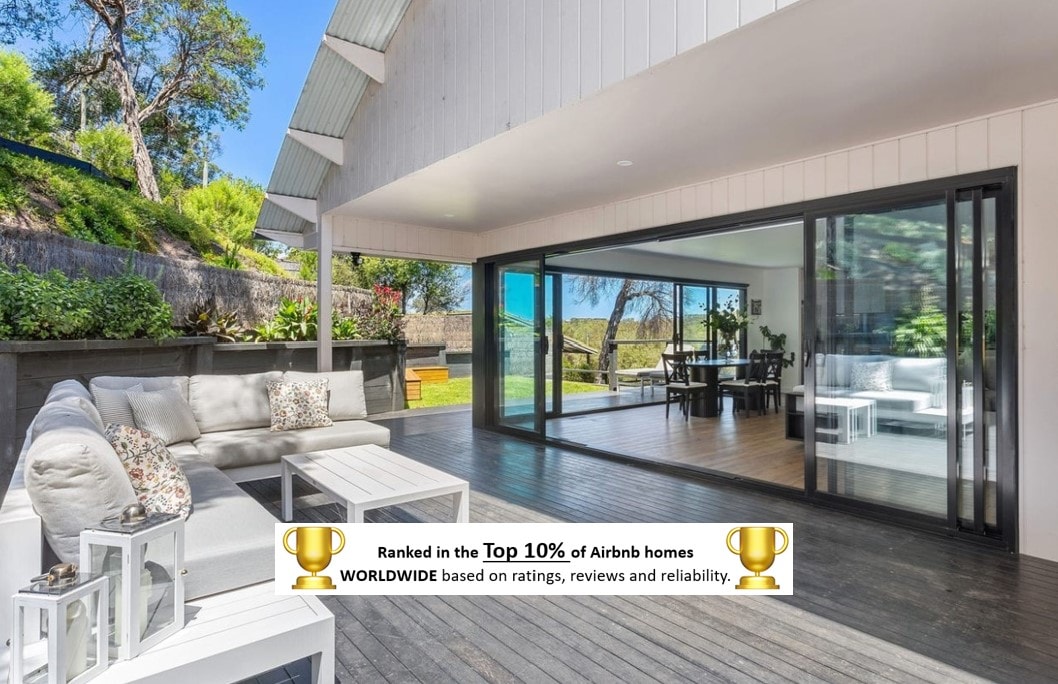 An inviting outdoor seating area is situated on a wooden deck, featuring plush seating and decorative pillows. Large sliding glass doors lead to an indoor dining space, enhancing the connection between indoor and outdoor living, while lush greenery surrounds the property.
