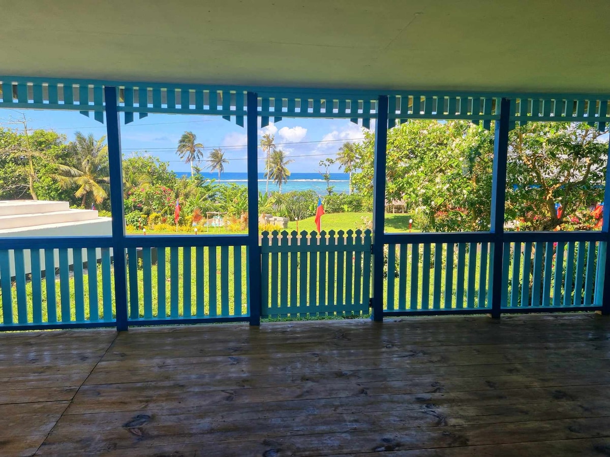 A spacious covered balcony is outlined by vibrant blue railings. Lush greenery fills the view, with palm trees swaying in the breeze. Beyond the garden, the ocean glimmers under sunlight, inviting relaxation and tranquility.
