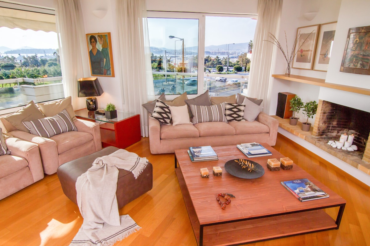 A spacious living room is filled with natural light from large windows overlooking the Aegean Sea. Two comfortable sofas adorned with decorative pillows sit adjacent to a stylish coffee table, complemented by warm wooden flooring and subtle artwork on the walls.
