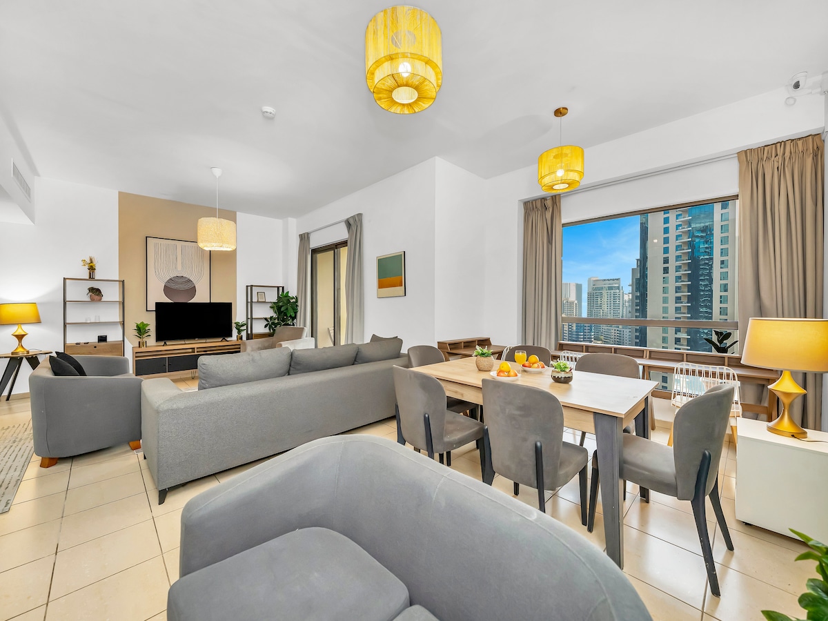 An open living area features a modern design with a large sectional sofa and a dining table. Natural light floods the space through expansive windows adorned with soft curtains. Decorative elements include indoor plants and stylish light fixtures, creating a comfortable atmosphere.