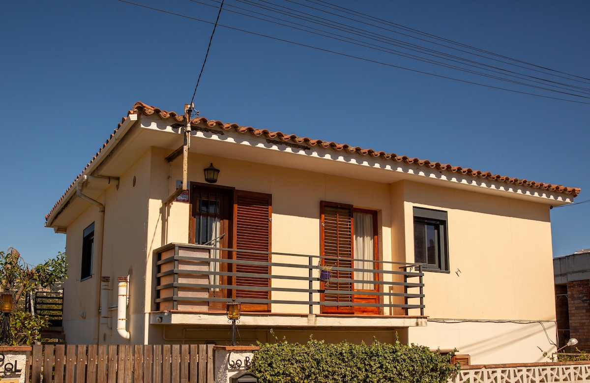 The exterior of a two-story house is shown, featuring a light-colored façade with a tiled roof. A balcony with wooden railings extends from the upper floor, complemented by several windows that allow natural light. The surrounding area includes greenery and a wooden fence.