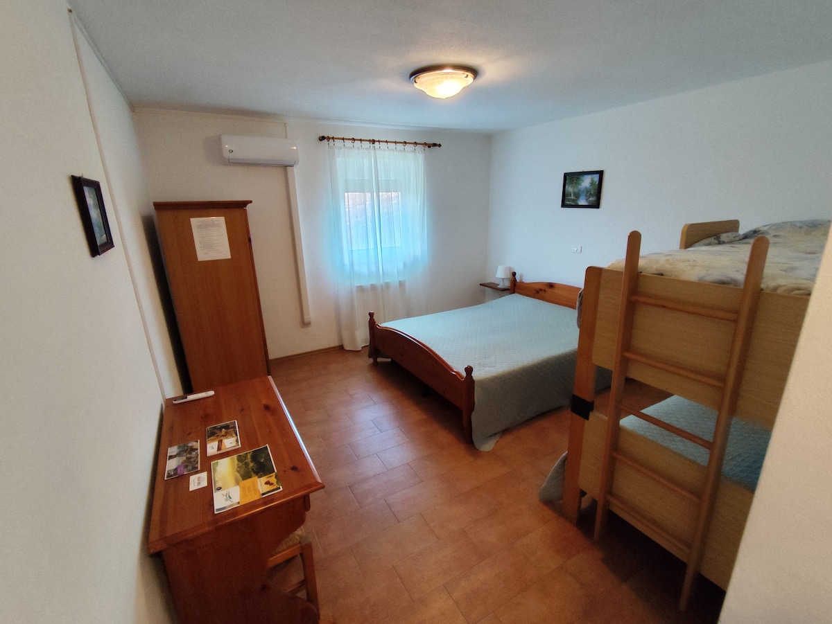 A well-lit family room features a spacious bed and a wooden bunk bed. A small desk with tourism brochures is positioned beside the bed, and a window is dressed with light curtains, allowing natural light to brighten the area. An air conditioning unit is mounted on the wall.
