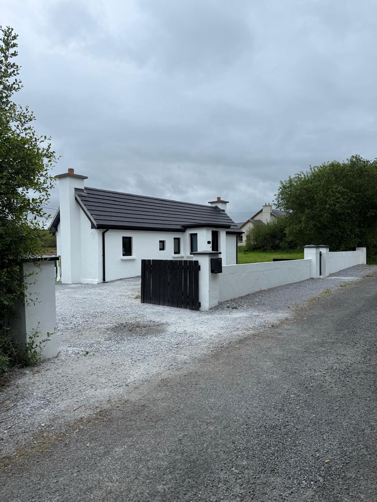 A modern cottage is visible from the gravel driveway, featuring a white exterior and a slate roof. The front yard is bordered by a low wall. Surrounding greenery enhances the cottage's peaceful setting, with a dirt road leading towards it.