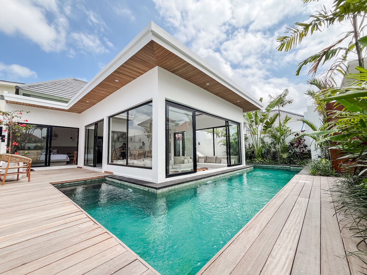The villa features a modern architectural design with expansive glass doors that invite natural light. A private pool is visible, surrounded by a wooden deck and tropical landscaping, creating a serene outdoor space for relaxation.