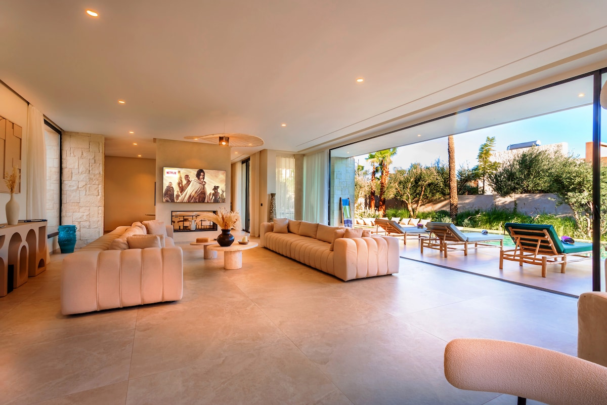 A spacious living area features a soft, neutral-colored sofa oriented towards a large wall with art. Expansive glass doors allow natural light to fill the room while providing views of the outdoor pool and garden area, with seating positioned for relaxation nearby.