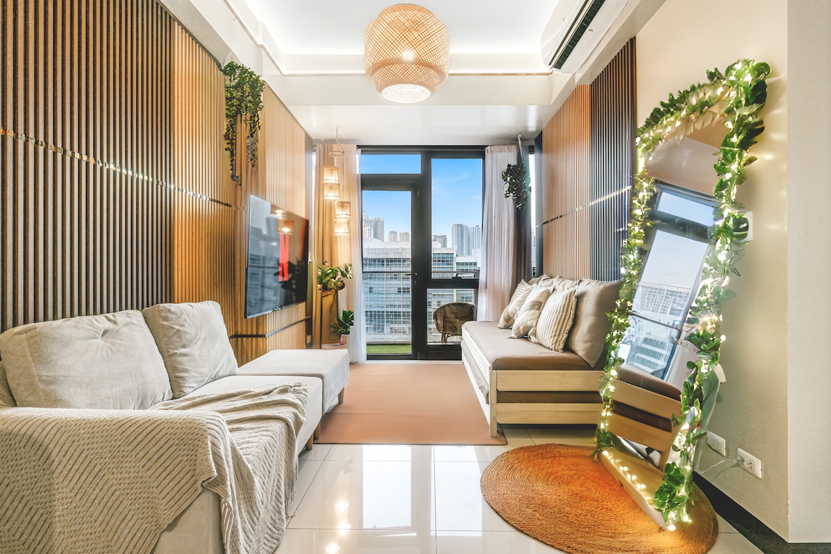 A cozy living area features warm wood accents and soft textures. A spacious couch sits beside a sleek TV mounted on the wall. Natural light floods through the large window, which offers a view of the city skyline. Decorative plants and fairy lights enhance the inviting ambiance.