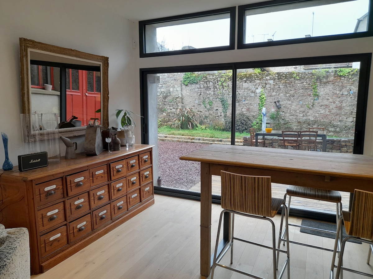 A bright dining area showcases a wooden table surrounded by bar stools, with large glass doors allowing views of a private outdoor space. A wooden sideboard with decorative items is positioned against the wall, adding a touch of warmth to the modern decor.