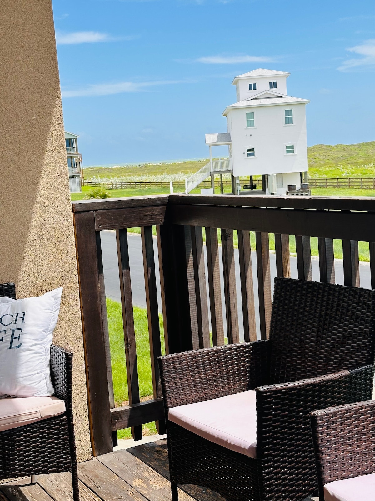 A private balcony features two wicker chairs with soft cushions, offering a relaxing seating area. A scenic view of a white building stands in the distance against a clear blue sky and vibrant green landscape, creating a peaceful outdoor space.