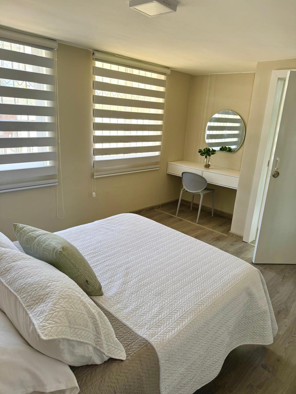 A bright bedroom features large windows covered with sleek horizontal blinds, allowing natural light to fill the space. A spacious bed is draped with a textured white coverlet. A small desk with a round mirror and a chair offers a cozy area for work or relaxation.
