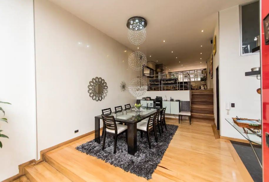 A spacious dining area features a large glass table surrounded by eight chairs. A cluster of decorative chandeliers provides soft lighting above. The wooden floor is complemented by a textured dark rug, contributing to an open and elegant atmosphere within the apartment.