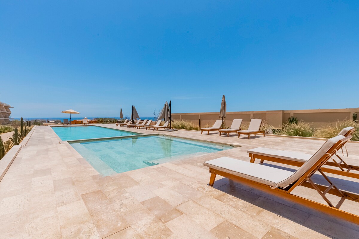 A spacious infinity pool is surrounded by a stone patio, featuring several lounge chairs arranged for relaxation. Umbrellas provide shade in designated areas. The horizon displays sweeping ocean views under bright blue skies, contributing to a tranquil atmosphere.