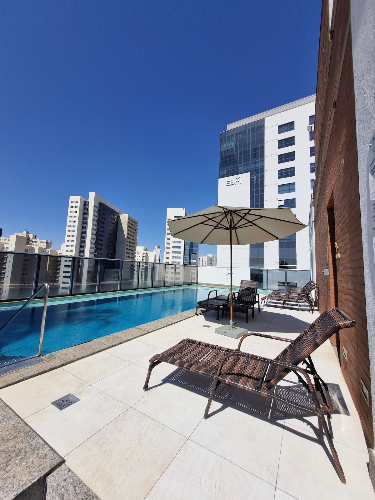 A rooftop pool area features a glistening blue pool bordered by smooth tiles. Several lounge chairs and a large umbrella provide shaded seating, while city buildings create a striking backdrop under a clear blue sky.