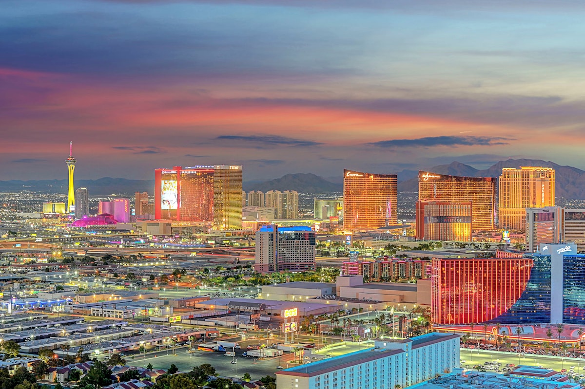 A panoramic view of the Las Vegas Strip at sunset, showcasing vibrant lights emanating from various high-rise buildings. The iconic Stratosphere stands tall on the left, while neighboring resorts reflect hues of orange and pink from the sky, creating a visually striking scene.