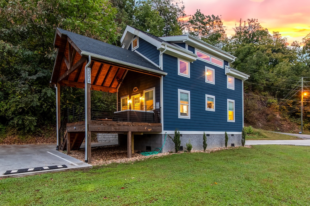 A modern blue cabin is set against a natural backdrop, featuring a spacious covered patio area. Large windows allow for ample natural light, while a manicured lawn leads to the driveway, providing convenient parking options.