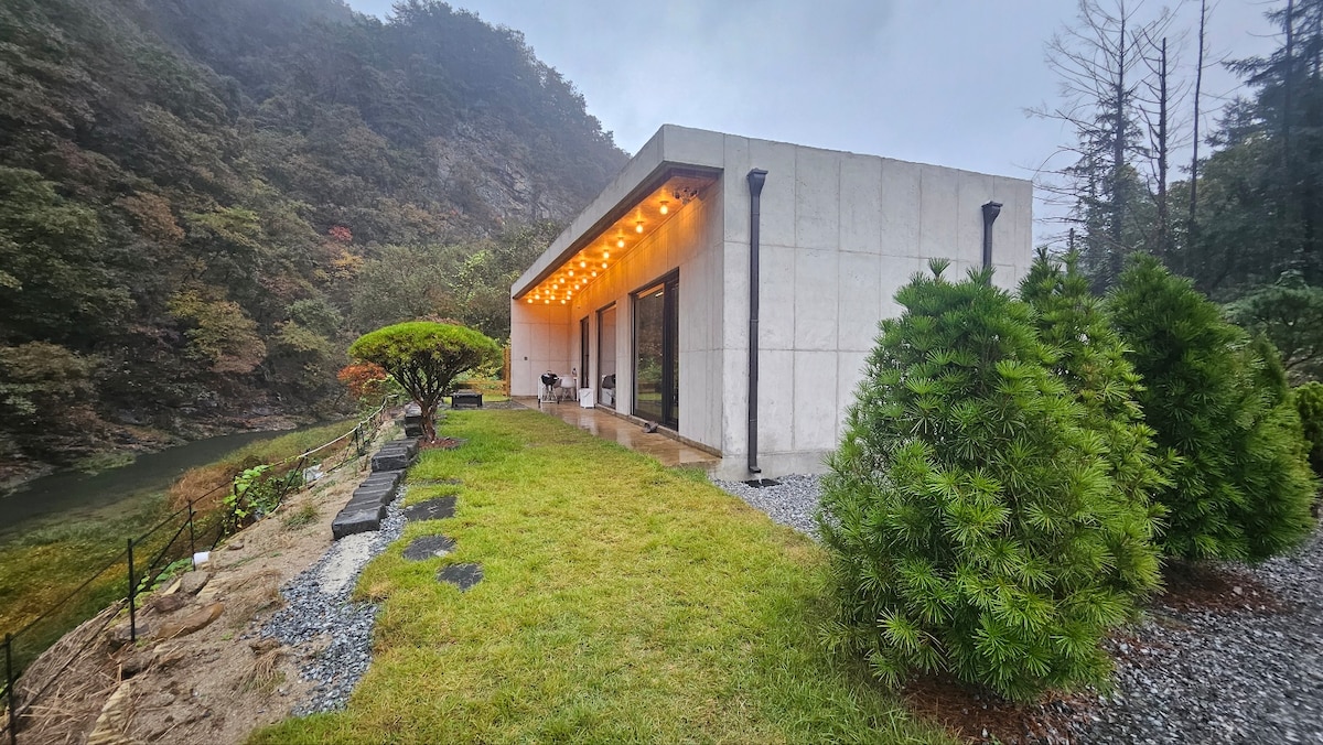 A modern single-story house is set against a lush, forested backdrop, featuring large glass doors that open to a grassy area. Soft lighting accentuates the exterior, while a winding stream can be seen nearby, surrounded by greenery and natural stones.