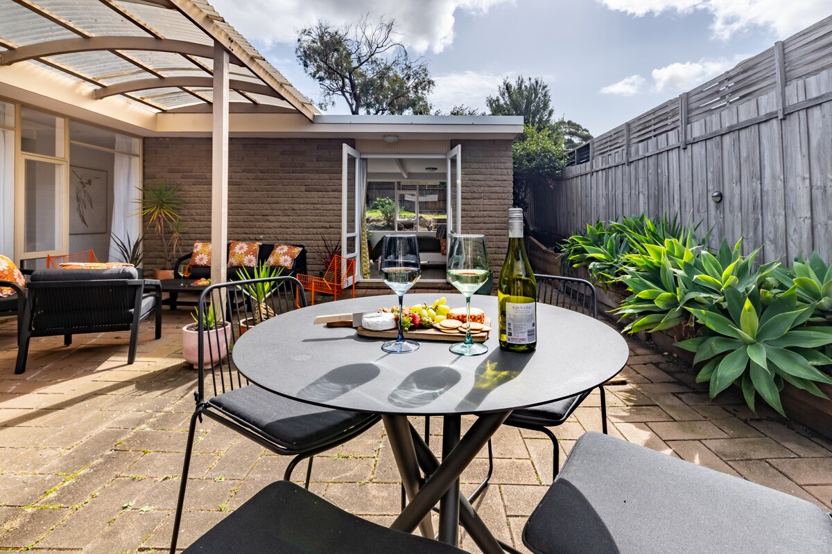 An outdoor seating area utilizes a round table set for two, featuring glasses of wine and a cheese platter. Colorful chairs surround the table, and a cozy lounge space is visible in the background. Lush greenery and a wooden fence create a serene atmosphere.
