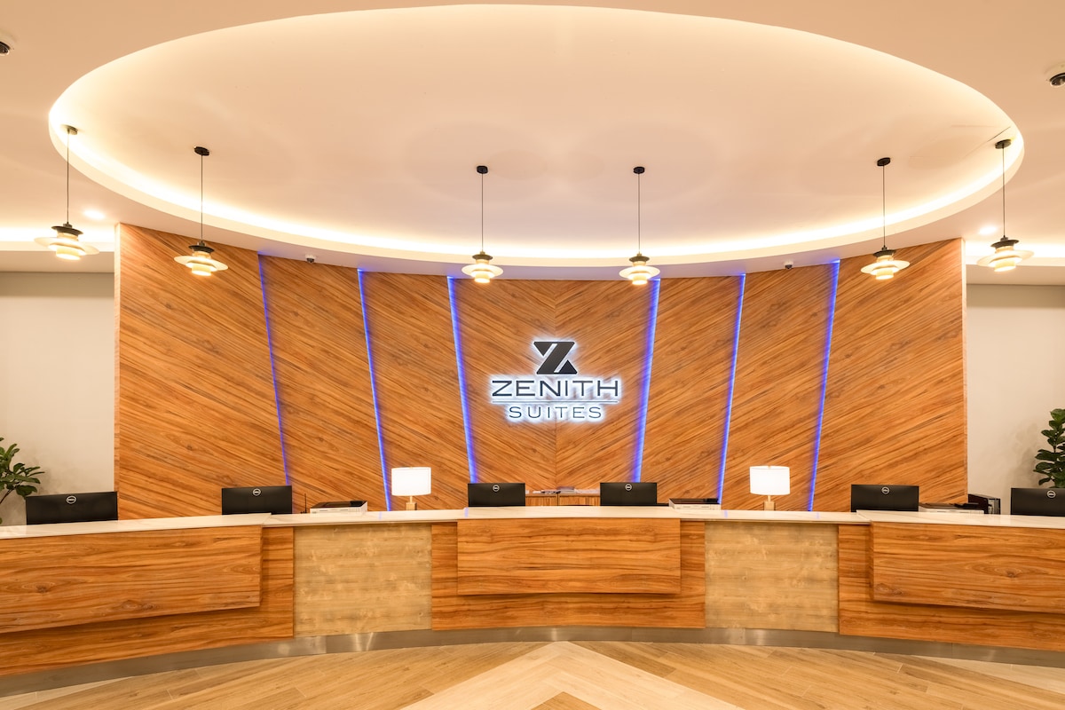 The reception area showcases a modern design with a wooden feature wall and illuminated accents. A curved countertop is complemented by several dark-colored check-in desks. Soft lighting is provided by pendant lights, creating a welcoming environment for guests.