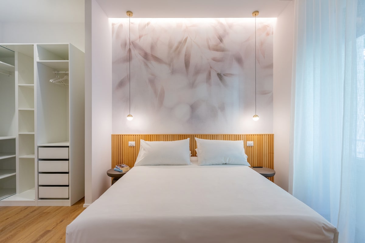 A serene bedroom features a spacious bed with crisp white linens, complemented by subtle pendant lights above. A soft, pastel-colored wall adds depth, while a built-in wardrobe provides organized storage options. Natural light filters through a sheer curtain, enhancing the calming ambiance of the space.