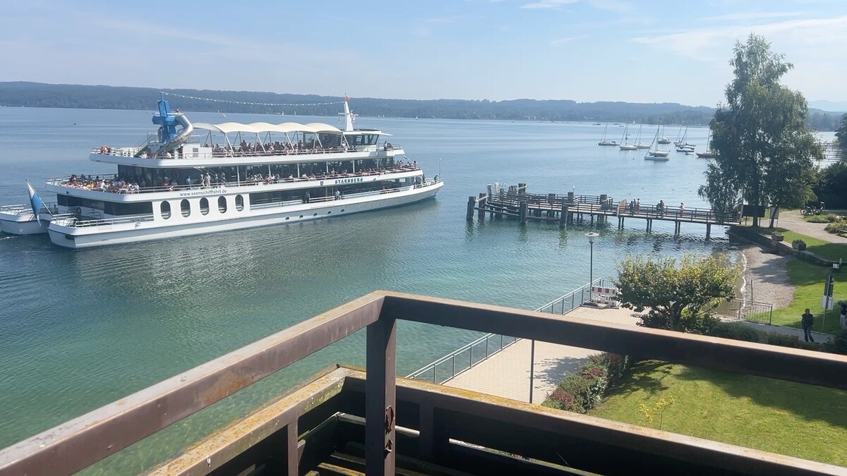 A scenic view from the balcony captures a boat cruising on Lake Starnberg. The image features a wooden railing in the foreground, with the calm, clear waters reflecting the sky and nearby trees. A pier with boats is visible to the right.