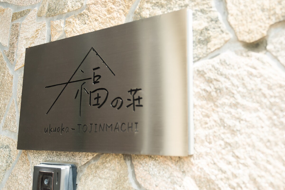 A metal sign is displayed, featuring the property's name '福田の庄' along with the text 'ukouka-TOJINMACHI'. The sign is mounted on a textured stone wall, adding a natural touch to the entrance.