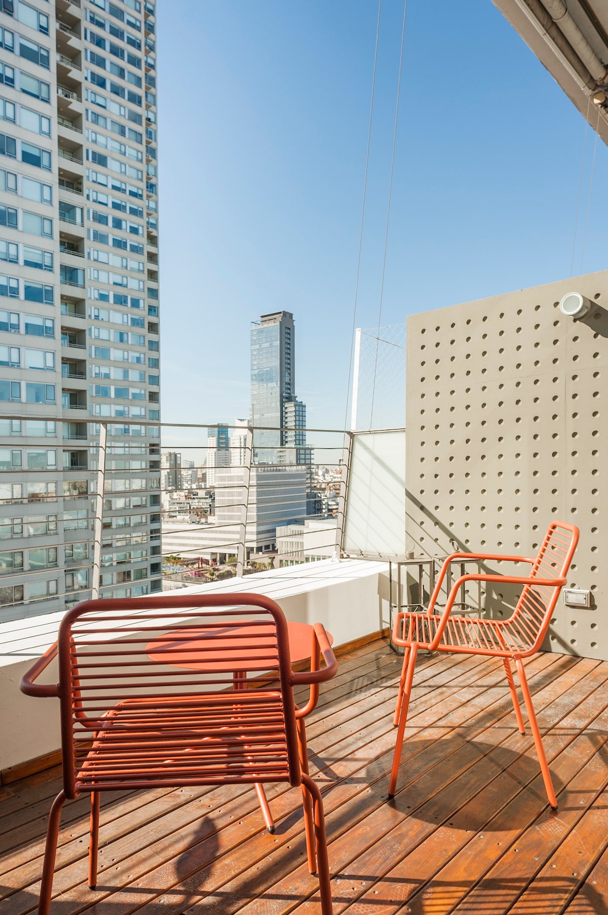 A private terrace reveals a panoramic view of the city, featuring two red outdoor chairs on a wooden deck. The surrounding high-rise buildings and a clear blue sky create a vibrant urban backdrop, ideal for relaxation and enjoying the scenery.